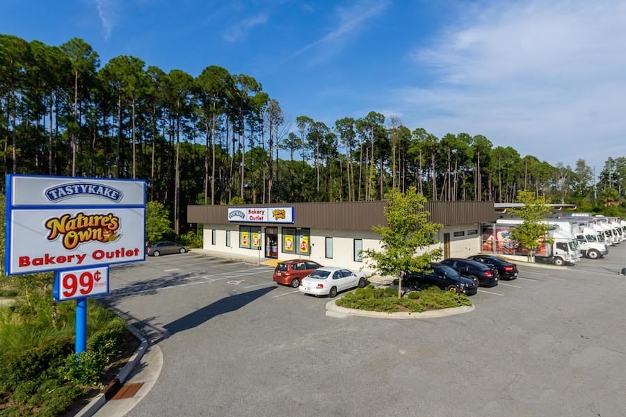 A small, single-story Flowers Foods bakery outlet surrounded by a parking lot with cars, located next to a wooded area under a clear sky.