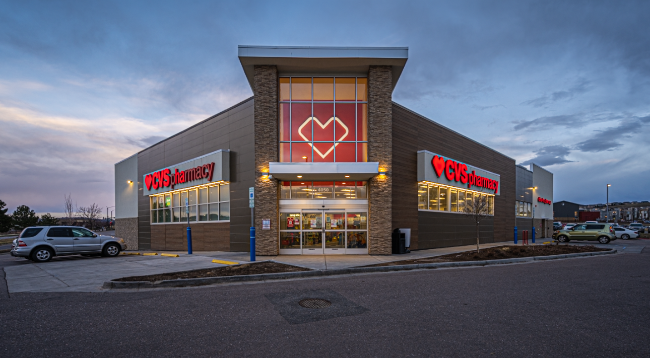Exterior of a recently sold CVS pharmacy store in Castle Rock, Colorado at twilight with illuminated signage and parked cars under a cloudy sky.