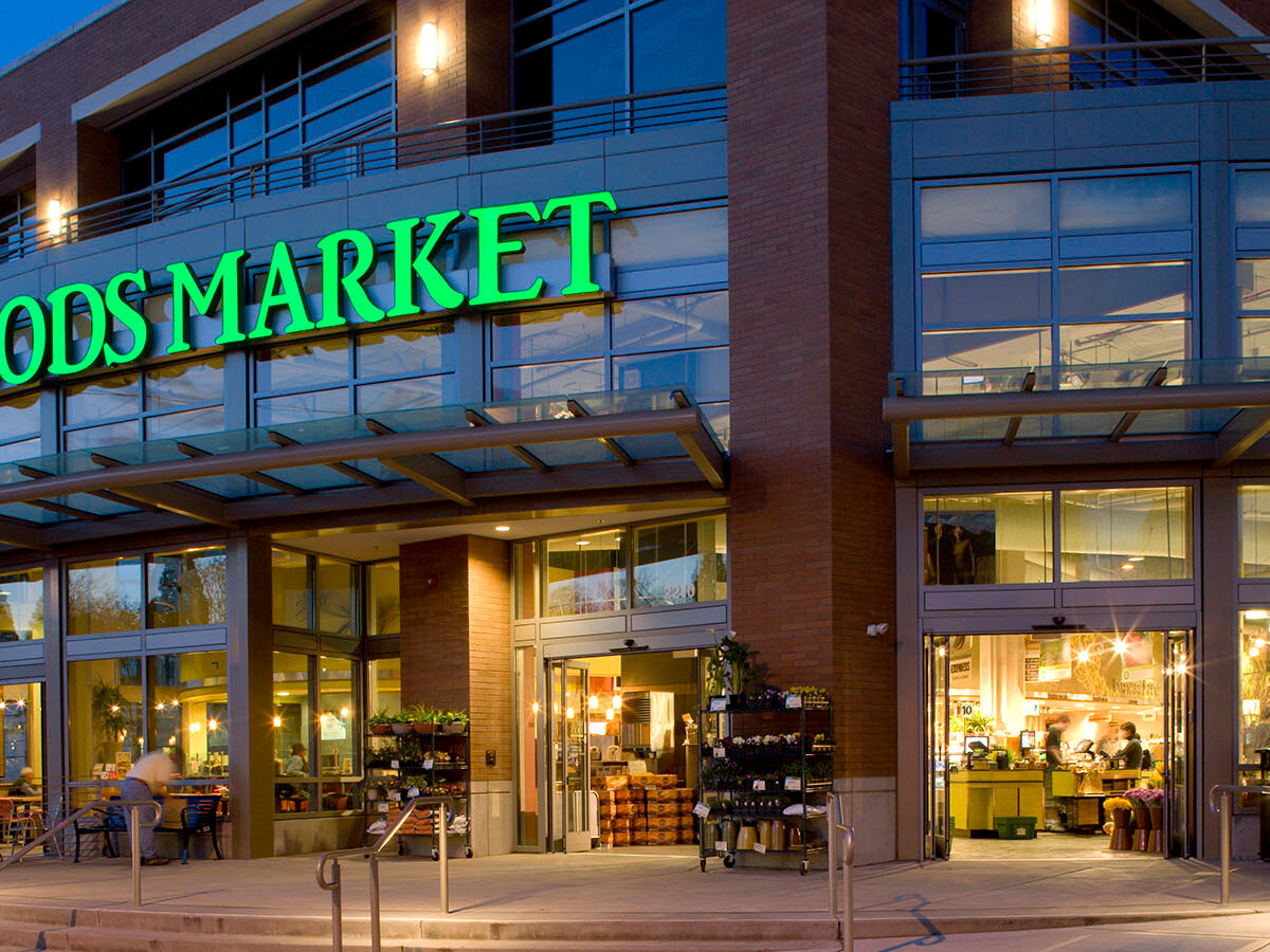 Exterior of a whole foods market at dusk with illuminated green signage, featuring a bicyclist at the entrance and visible interior lighting, exemplifying prime single tenant net leased investments nationwide.