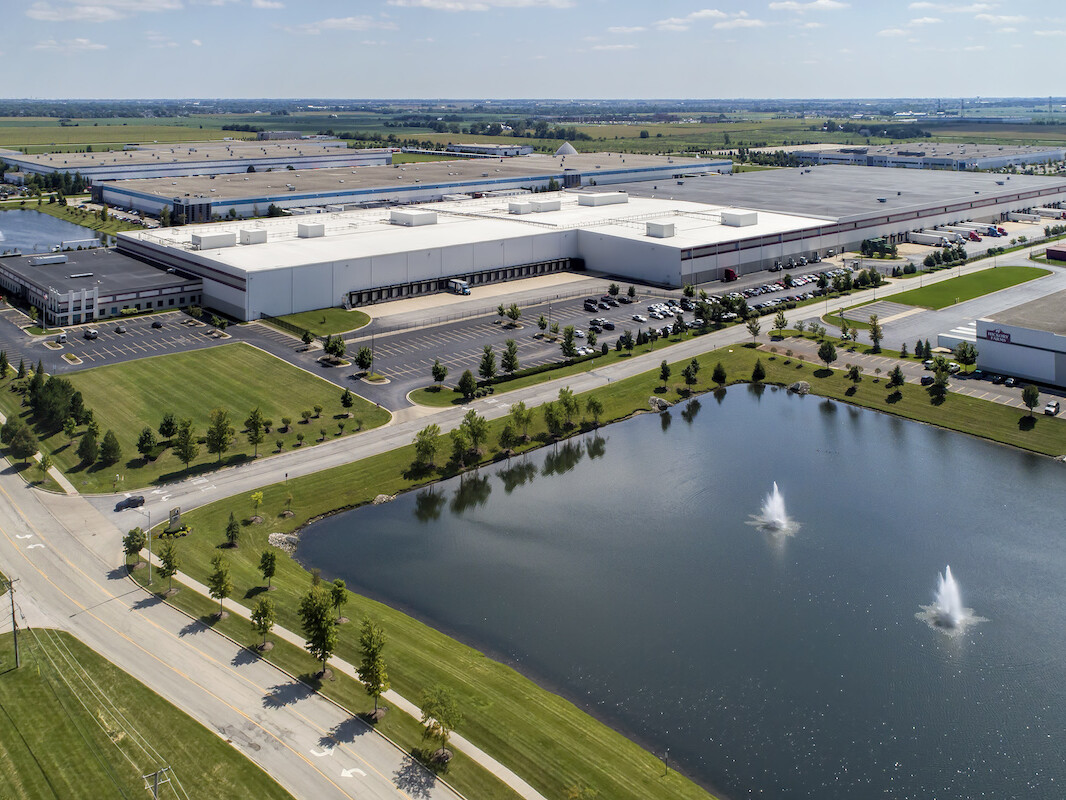 Aerial view of a sprawling Supervalu Inc industrial park with multiple warehouses, a parking lot, and a small lake with fountains, surrounded by greenery.