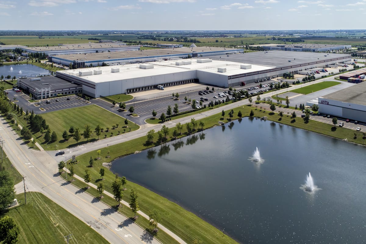Aerial view of a sprawling Supervalu Inc industrial park with multiple warehouses, a parking lot, and a small lake with fountains, surrounded by greenery.