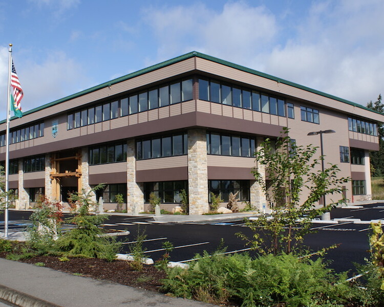 Modern two-story office building serving as a Washington State Parks facility, featuring a stone base and brown paneling, surrounded by a parking lot and green landscaping, under a clear sky.