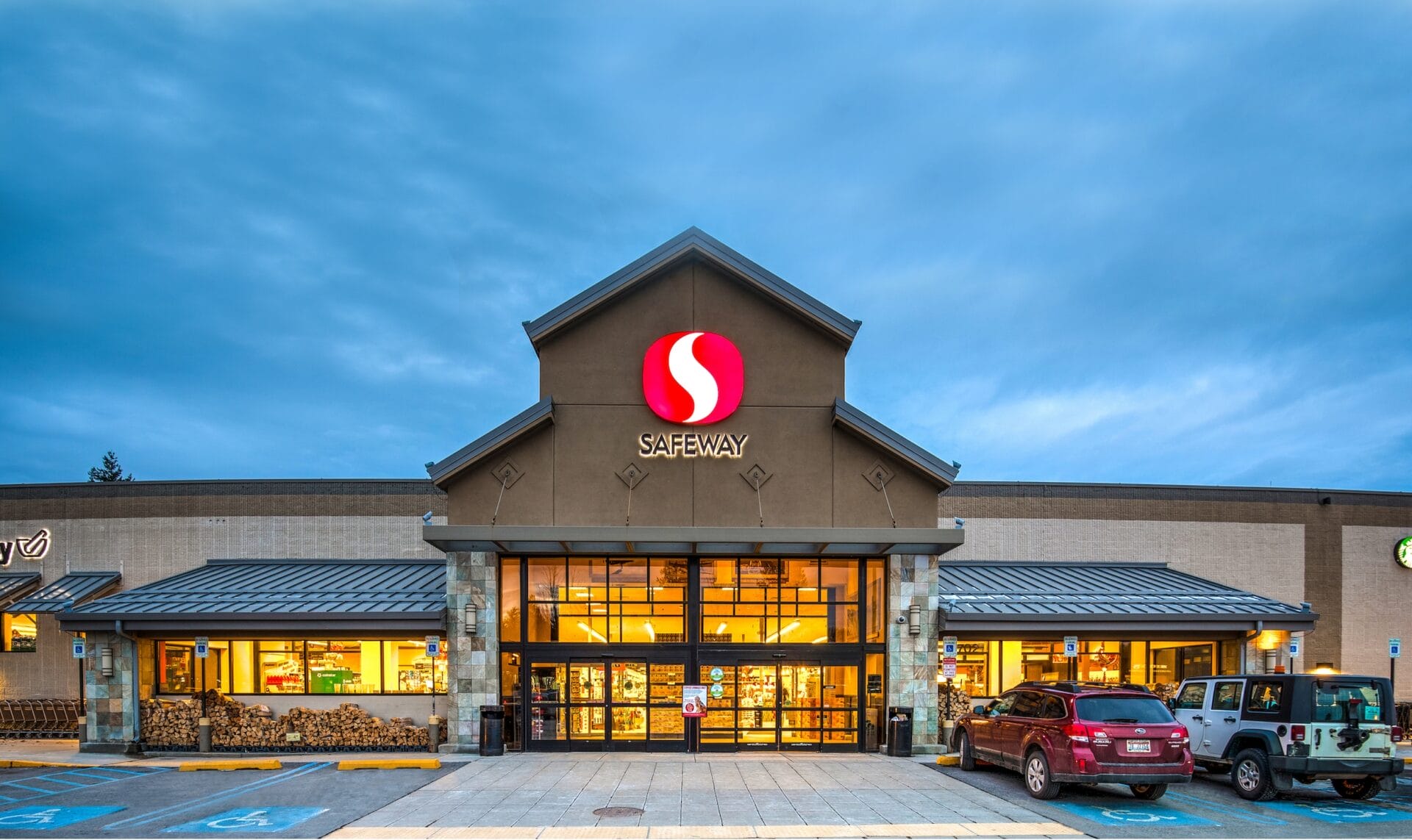 Exterior of a Safeway supermarket at dusk with illuminated signage and parked cars.