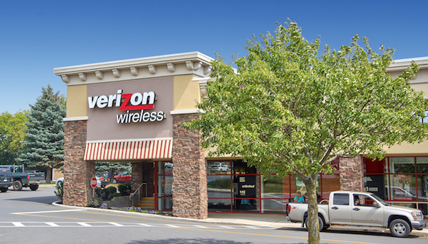 Exterior view of a recently sold Verizon Wireless store in a suburban shopping plaza with parked cars and trees under a clear sky.