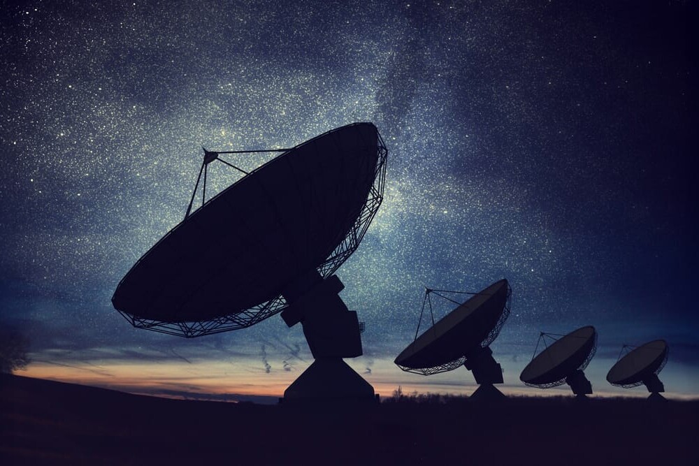 Four large radio telescopes, part of a major broadcasting facility, are aligned under a star-filled night sky, silhouetted against the fading light of dusk.