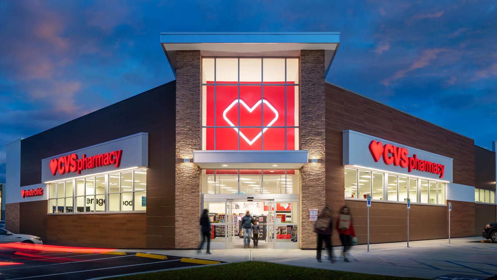 Exterior view of a recently sold CVS Pharmacy store at dusk with illuminated signage, large glass entrance, and several people entering and exiting.