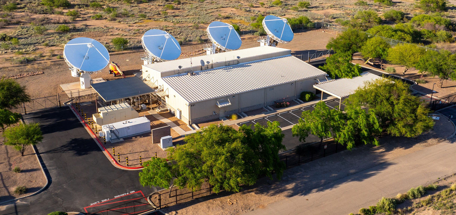 Aerial view of a fenced DIRECTV Data Center with four large satellite dishes on the roof, surrounded by desert vegetation and paved roads; this broadcasting facility was recently sold.