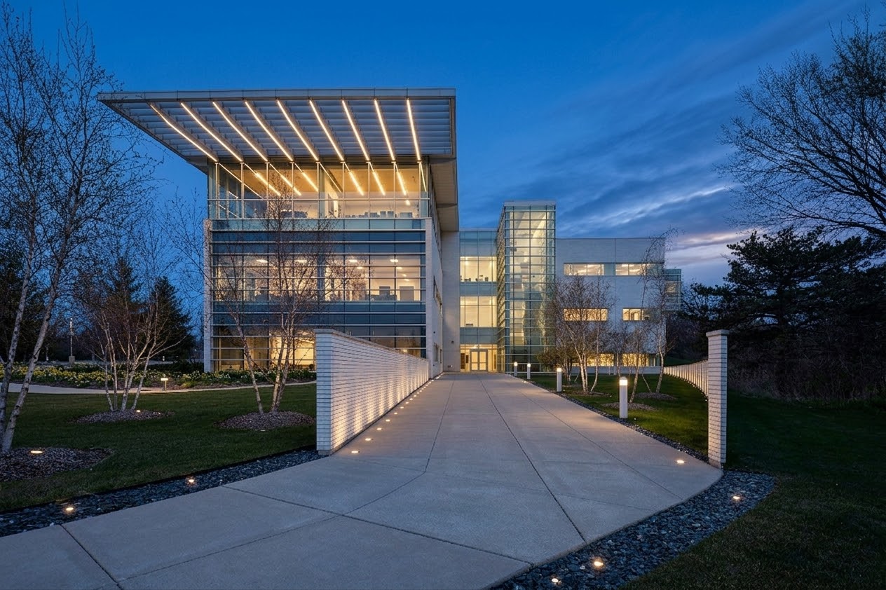 Modern glass office building with large overhanging roof, illuminated walkway, and landscaped grounds at dusk, scheduled for completion in September 2026.